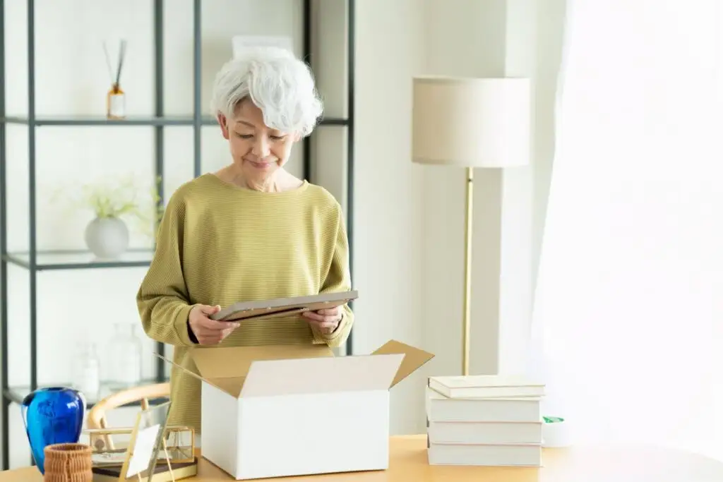 The Psychology Behind Why So Many Retirees Feel Happier After Simplifying Their Lives 2 A senior asian woman Downsizing After Retirement by packing belonging in a box to move to The Village at Duxbury in MA.